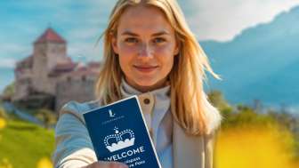 A woman holds up the WELCOME Experience Pass in front of Vaduz Castle in a field of flowers