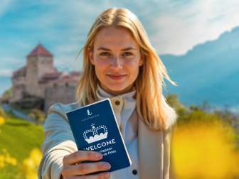 A woman holds up the WELCOME Experience Pass in front of Vaduz Castle in a field of flowers